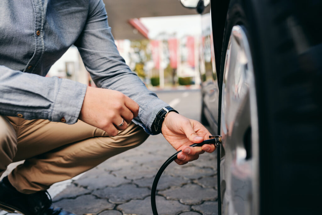 Homem abaixado próximo à roda de um carro, segurando uma mangueira de calibrador para pneus.